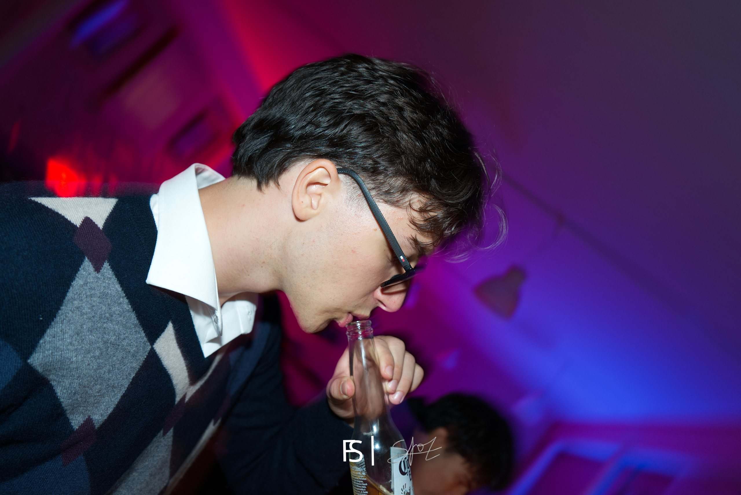Ragazzo con gli occhiali chino su una bottiglia, illuminato da luci magenta e blu